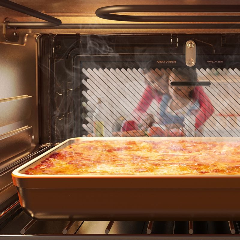 Foto vista de dentro do forno elétrico de bancada da Continental branco, modelo BC44B. Na prateleira, há um refratário retangular grande com lasanha fumegante. Do lado de fora do forno, uma menina corta um alimento com a ajuda de uma mulher.
