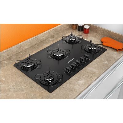 Foto-tirada-de-cima-pra-baixo-levemente-na-diagonal-do-cooktop-5-bocas-Continental-a-gas-de-vidro-temperado-preto-modelo-KC5GP-–-bivolt-instalado-em-uma-bancada-com-marmore-bege.-Sao-2-bocas-na-lateral-esquerda-uma-maior-no-meio-e-2-na-lateral-direita.-Todas-elas-com-grades-individuais.-Na-parte-da-frente-da-boca-do-meio-ficam-os-botoes-dos-queimadores.-Ha-utensilios-de-cozinha-ao-redor-do-produto-como-luva-termica-para-forno-e-fogao-e-potes-de-tempero.