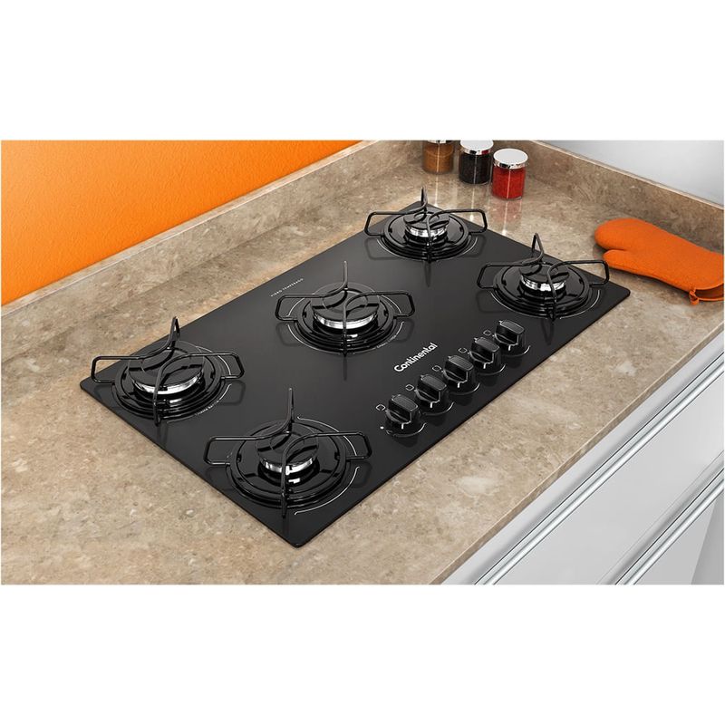 Foto tirada de cima pra baixo levemente na diagonal do cooktop 5 bocas Continental a gás de vidro temperado preto, modelo KC5GP – bivolt instalado em uma bancada com mármore bege. São 2 bocas na lateral esquerda, uma maior no meio e 2 na lateral direita. Todas elas com grades individuais. Na parte da frente da boca do meio, ficam os botões dos queimadores. Há utensílios de cozinha ao redor do produto, como luva térmica para forno e fogão e potes de tempero.