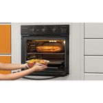 Foto do forno elétrico de embutir da Continental com grill e timer, na cor preta, modelo OC8EP, instalado em um móvel cinza. Mãos femininas usam um pano para tirar um refratário redondo da parte inferior do forno. Acima da prateleira há um refratário retangular com lasanha.