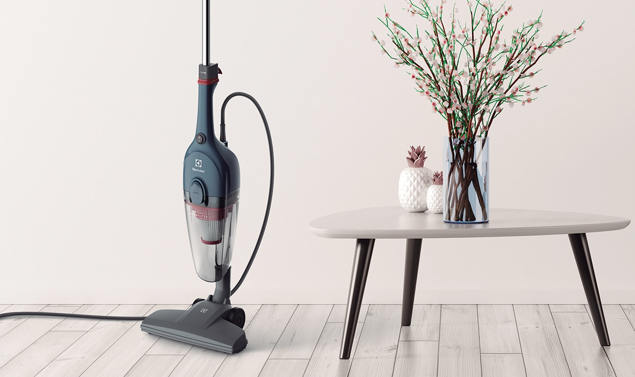 Aspirador vertical com fio Electrolux na cor azul  posicionado sobre um piso de madeira clara ao lado de uma mesa baixa com vasos decorativos numa foto ambientada 